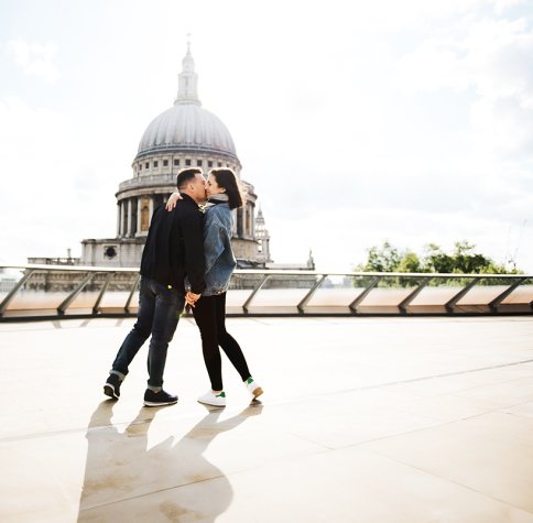 A young white man and white woman kissing on a bridge in front of St Paul's Cathedral.